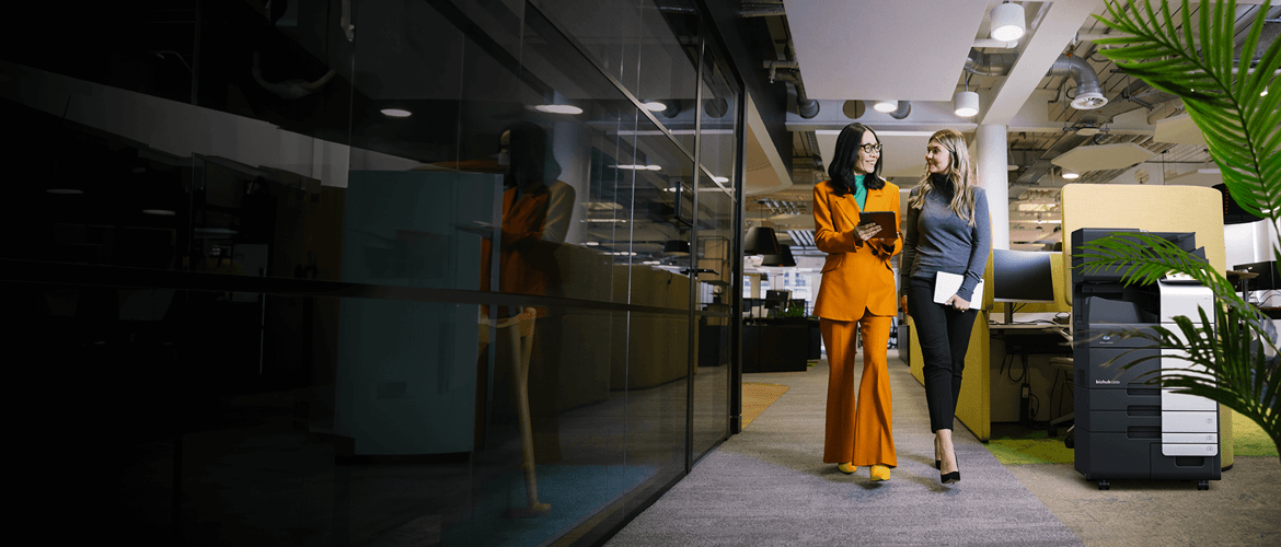 Two Businesswomen walking in a modern office past a A3 copier to represent Konica Minolta Business Solutions