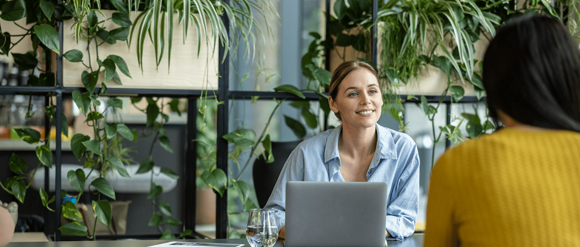 a women sitting down with a laptop looking at another person talking and looking happy