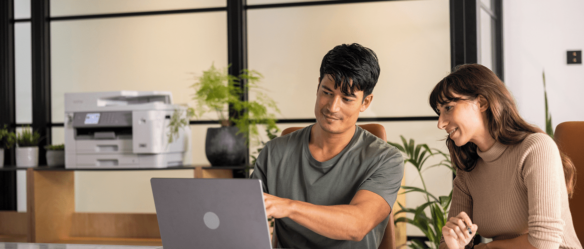 A man and woman at home sitting at a desk looking at a laptop with a printer and plants in the background to represent sustainability policy