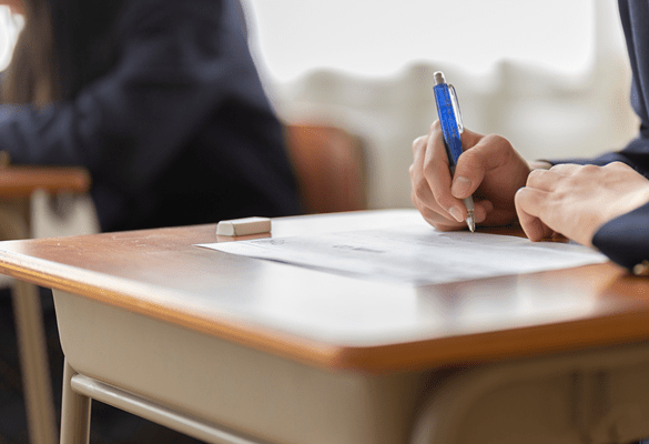 A student at a desk completing a test at school. The person is out of frame and only shows their hands holding a pen. This accompanies information about the results of Queen Charlotte College choosing Brother's Managed Print Services